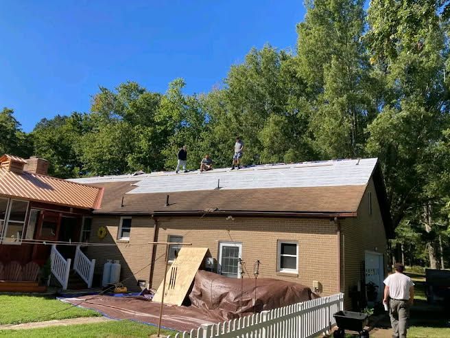 Workers on a roof with visible construction in progress. Brick building, green trees, and clear sky.