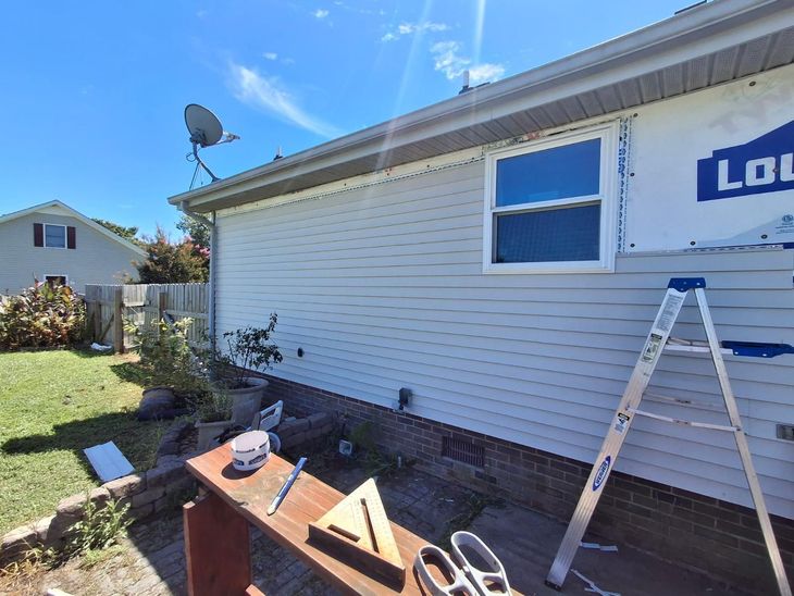 Exterior of a house with light siding and a new window. A ladder, tools, and a Lowe's sign are nearby, under a blue sky.