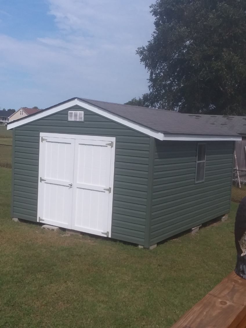 Green shed with white double doors, small window, and dark gray roof on a grassy lawn.