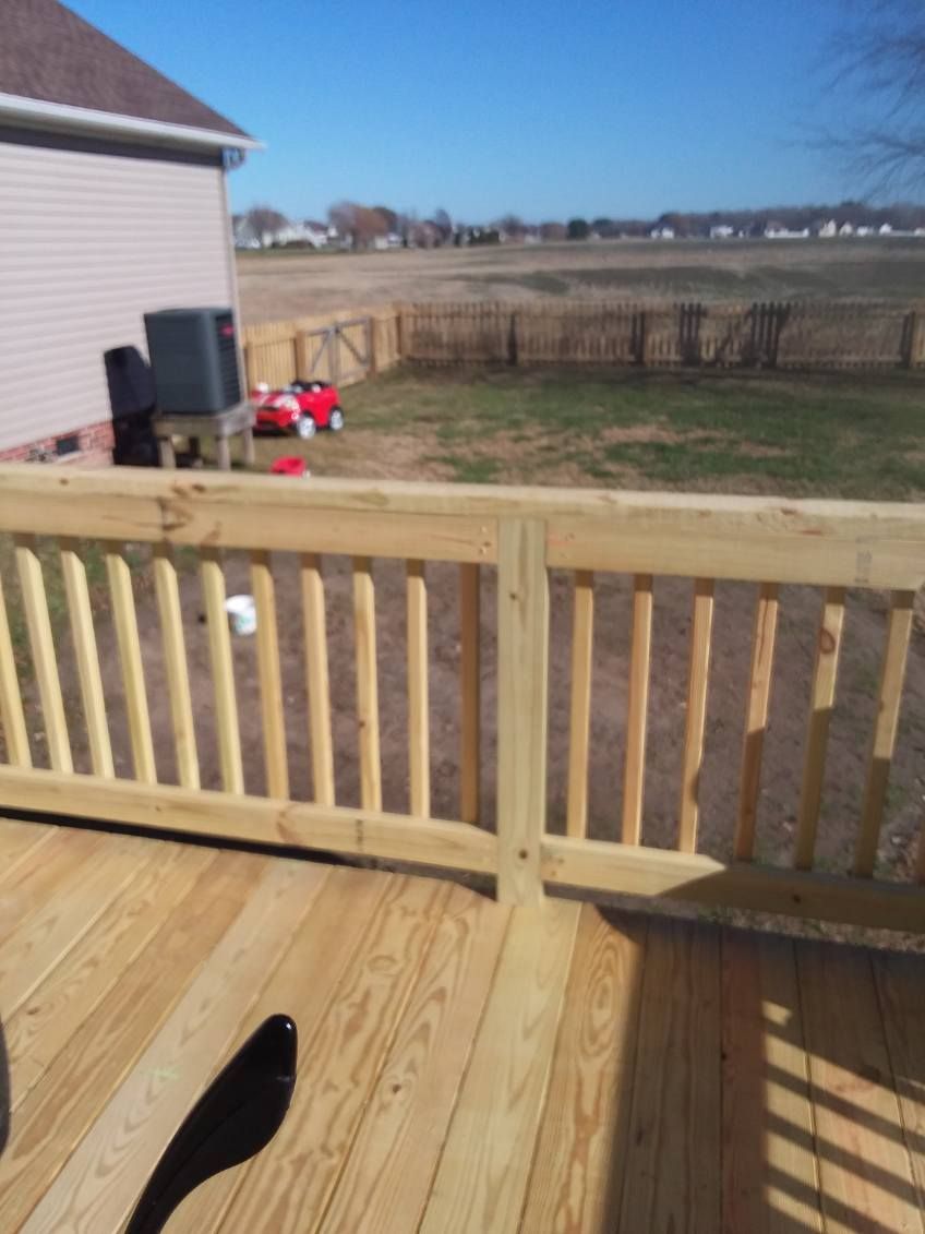 Wooden deck with railing, house exterior, and fenced backyard under a bright blue sky.