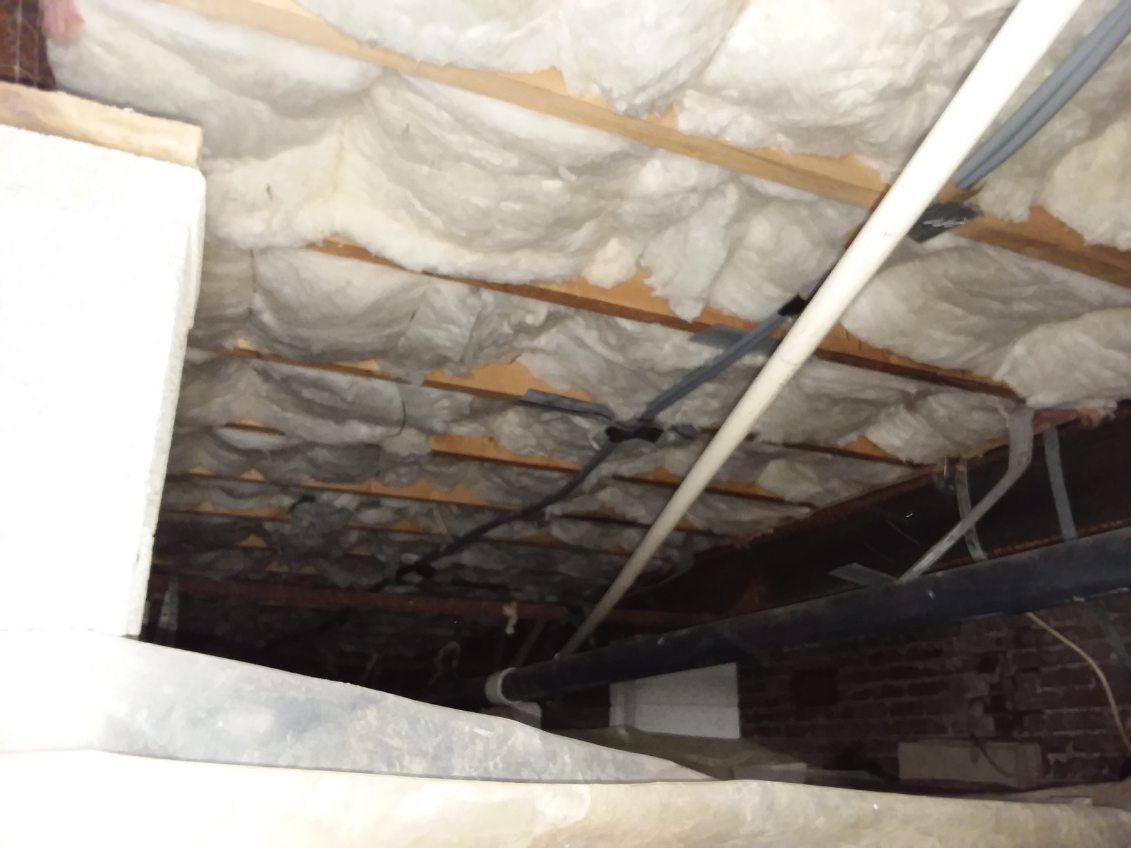 Crawlspace view: wood beams, insulation, pipes, brick wall. Dark, enclosed space.