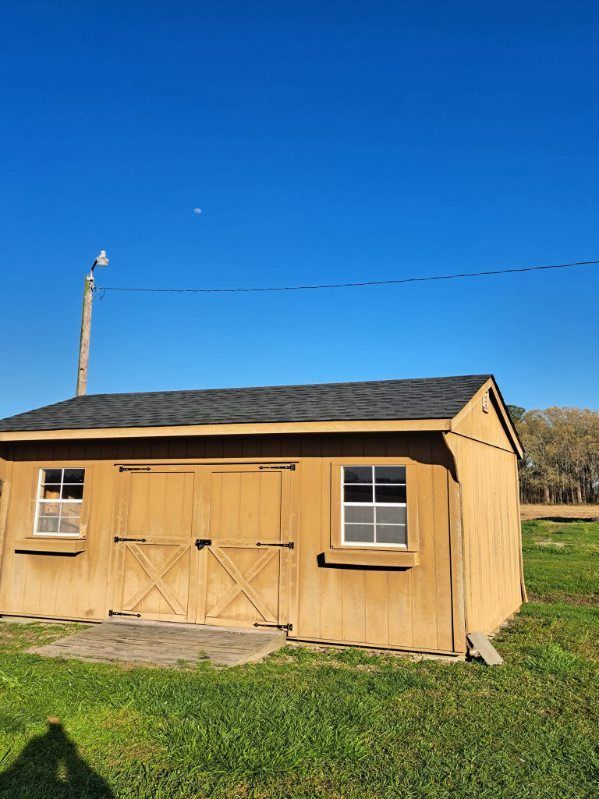 Tan wooden shed with black roof, two windows, and double doors on a grassy field, under a blue sky.