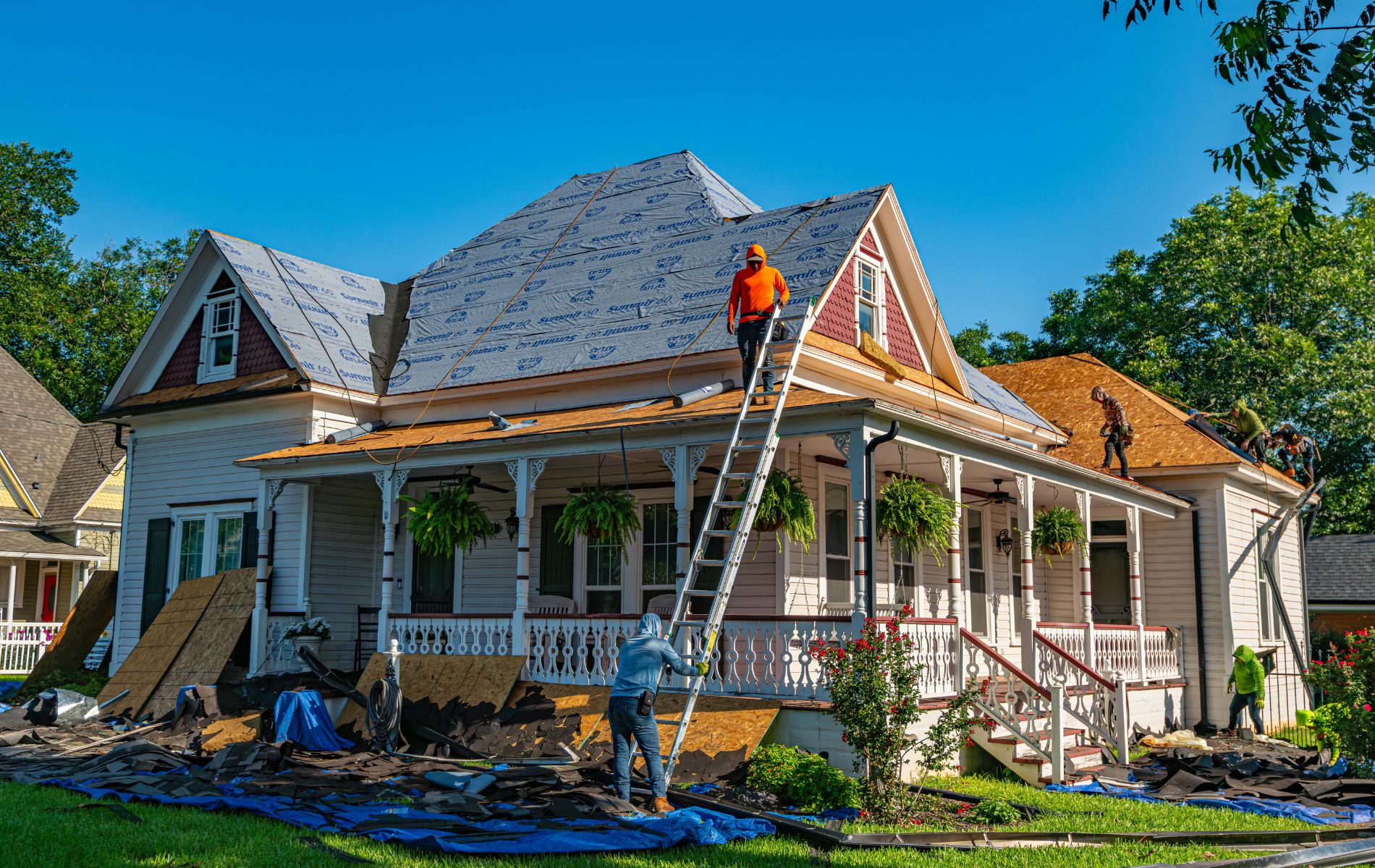 House undergoing roof replacement, workers on ladder, porch with hanging plants.