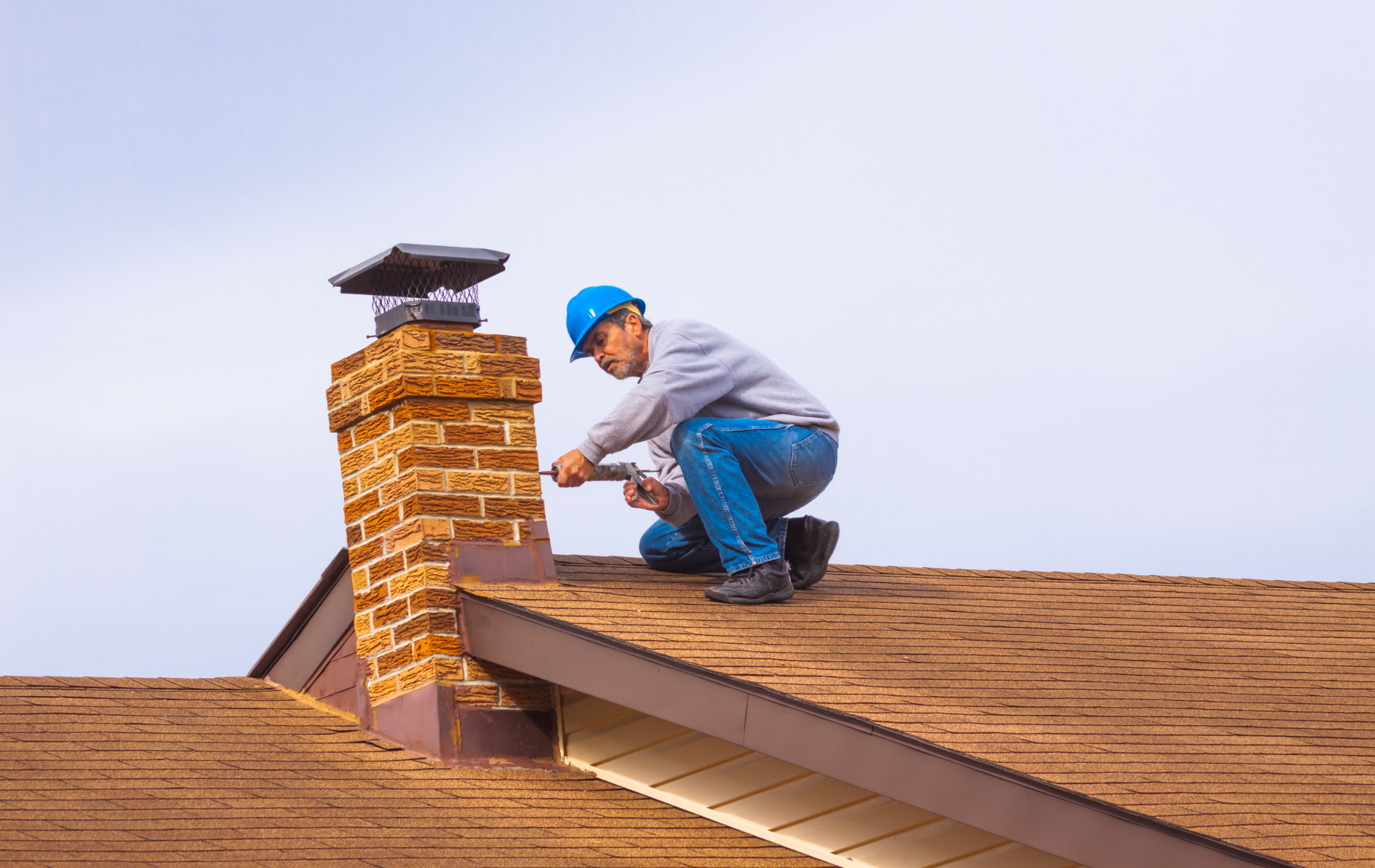 Person in blue hard hat on a roof repairs a brick chimney.
