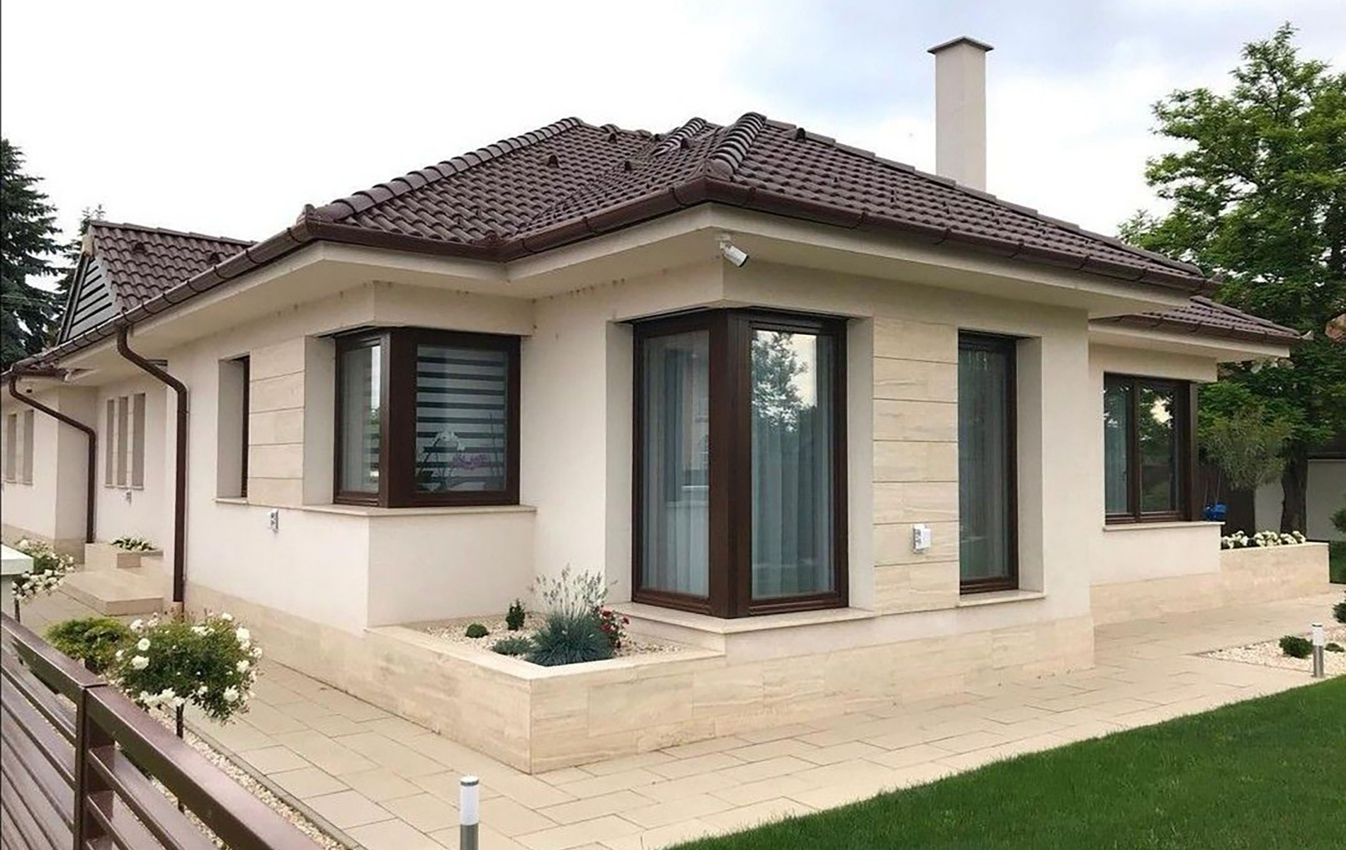 Beige modern house with brown roof and window frames, greenery around.