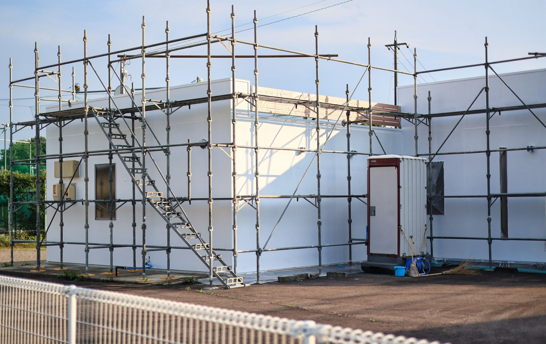 Building under construction, surrounded by scaffolding. White exterior, small door, metal stair.