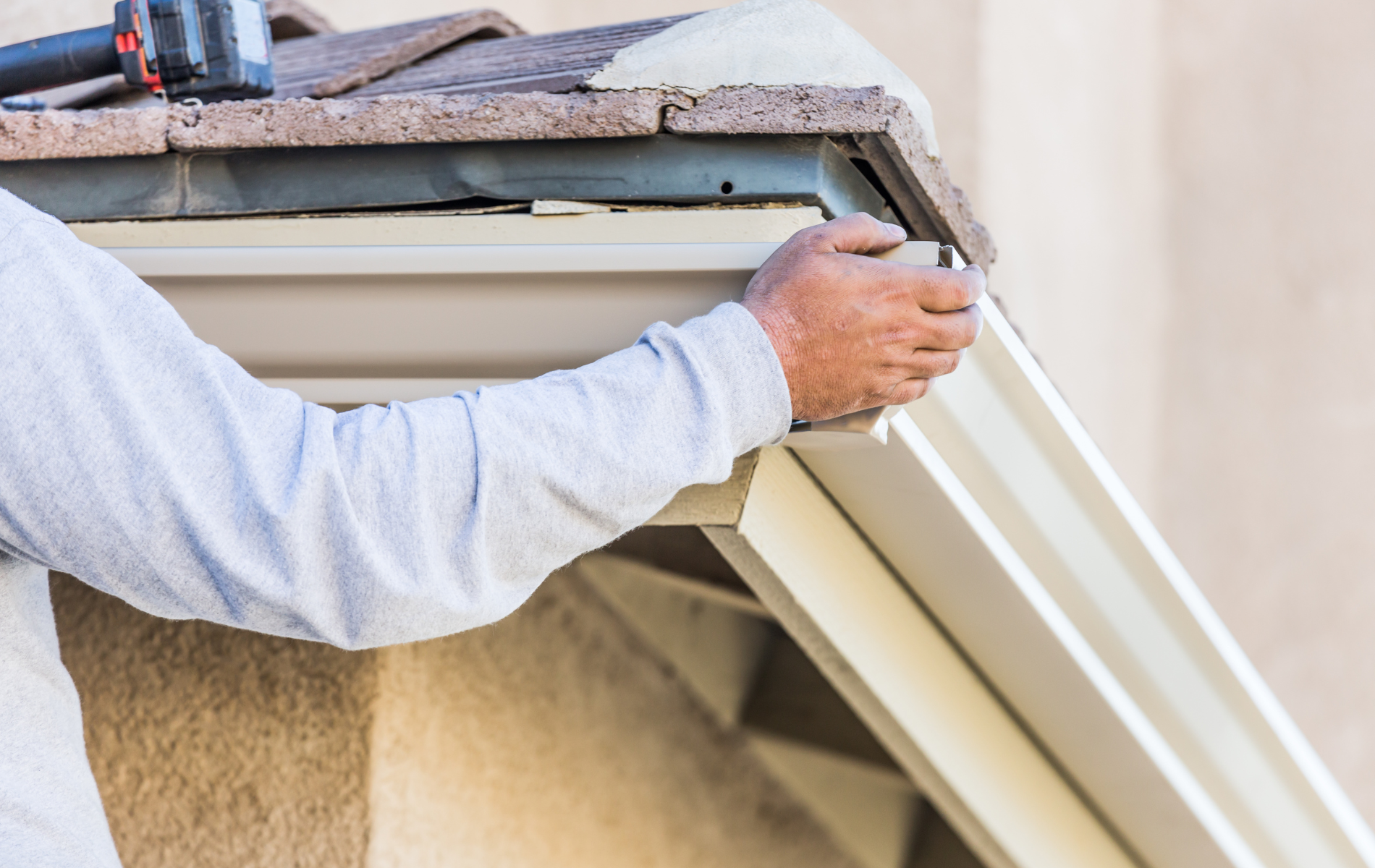 Person installing beige rain gutter on a house, using a power drill.