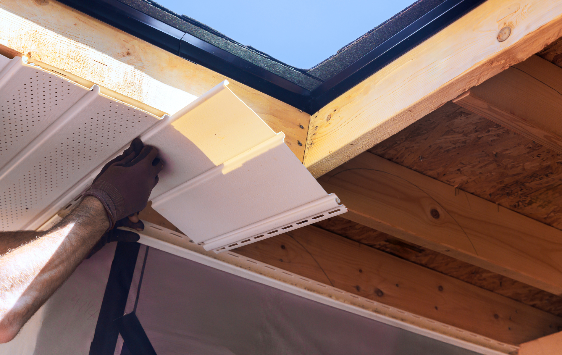 A person installing white vinyl siding around a skylight. Wooden structure, blue sky.