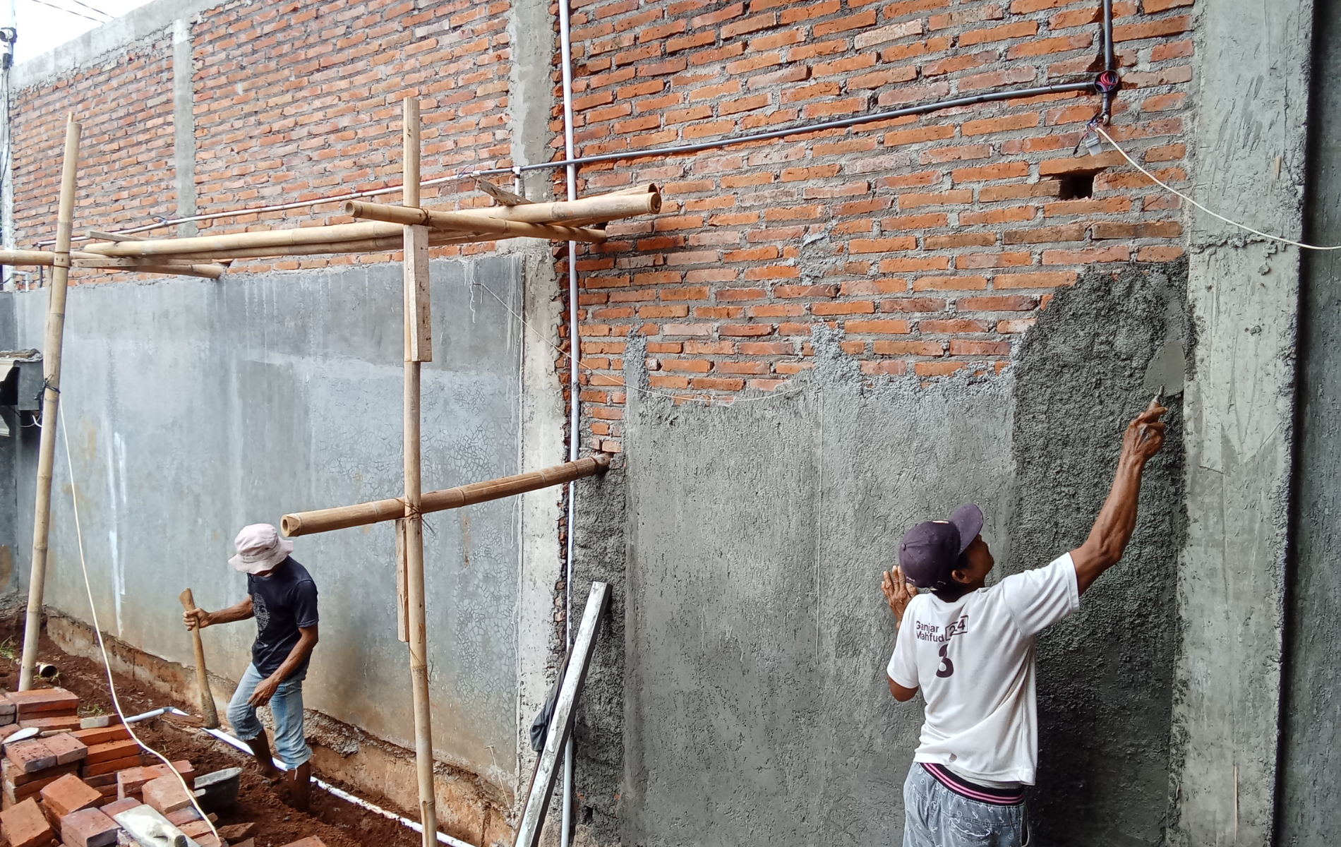 Construction workers plastering a brick wall. One worker applies plaster; the other digs in the dirt.