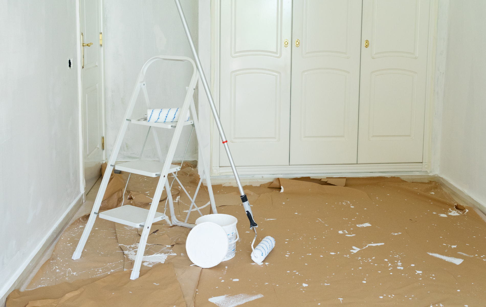 Room with painting supplies: stepladder, paint can, roller, drop cloth on floor.