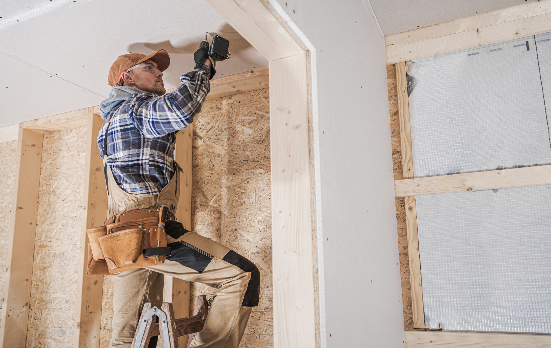 Man on stepladder installing drywall in a wooden framed interior. He's wearing work clothes and using a drill.