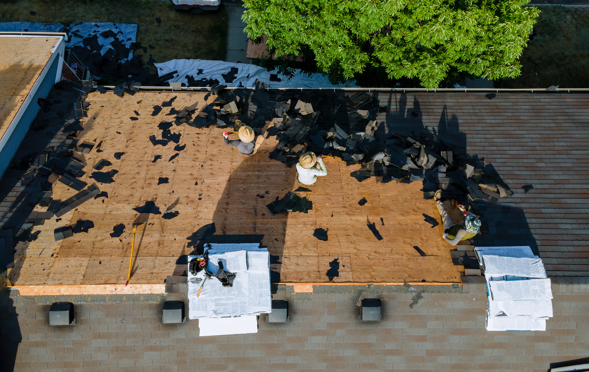 Aerial view of a roof being repaired by workers removing old shingles. Brown, black, and white materials are visible.