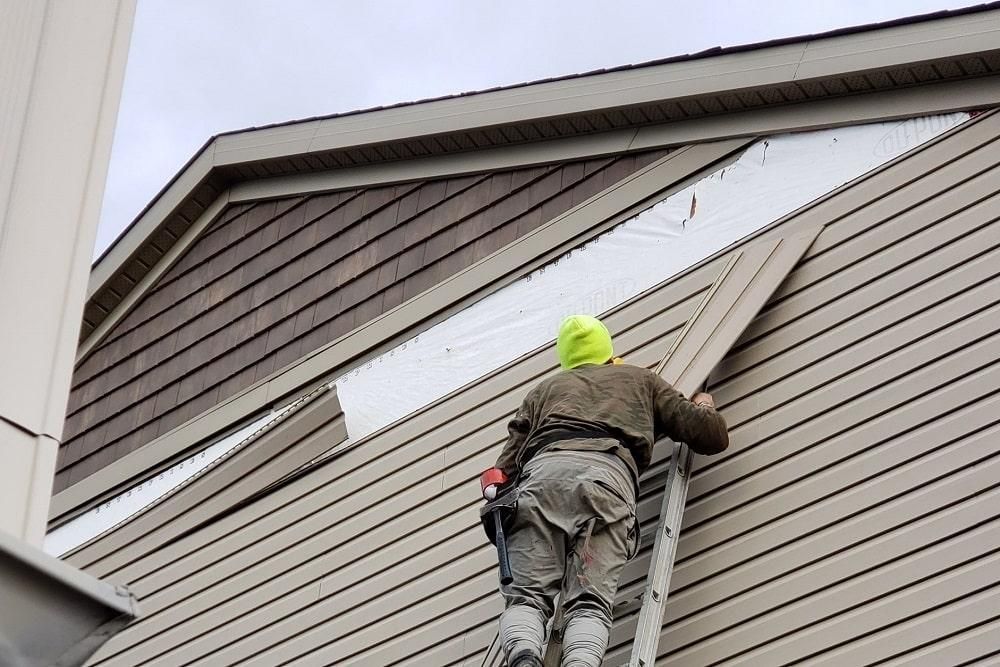 Person on ladder installing siding on a house, silver insulation visible. Overcast day.