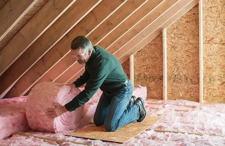 Man installing pink insulation in an attic. He kneels on a plywood board, holding a roll of insulation.