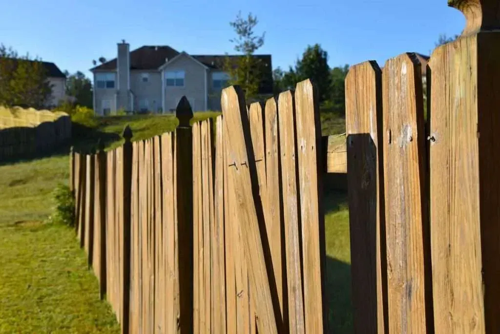Wooden fence in the foreground with a light-colored house visible in the background on a sunny day.