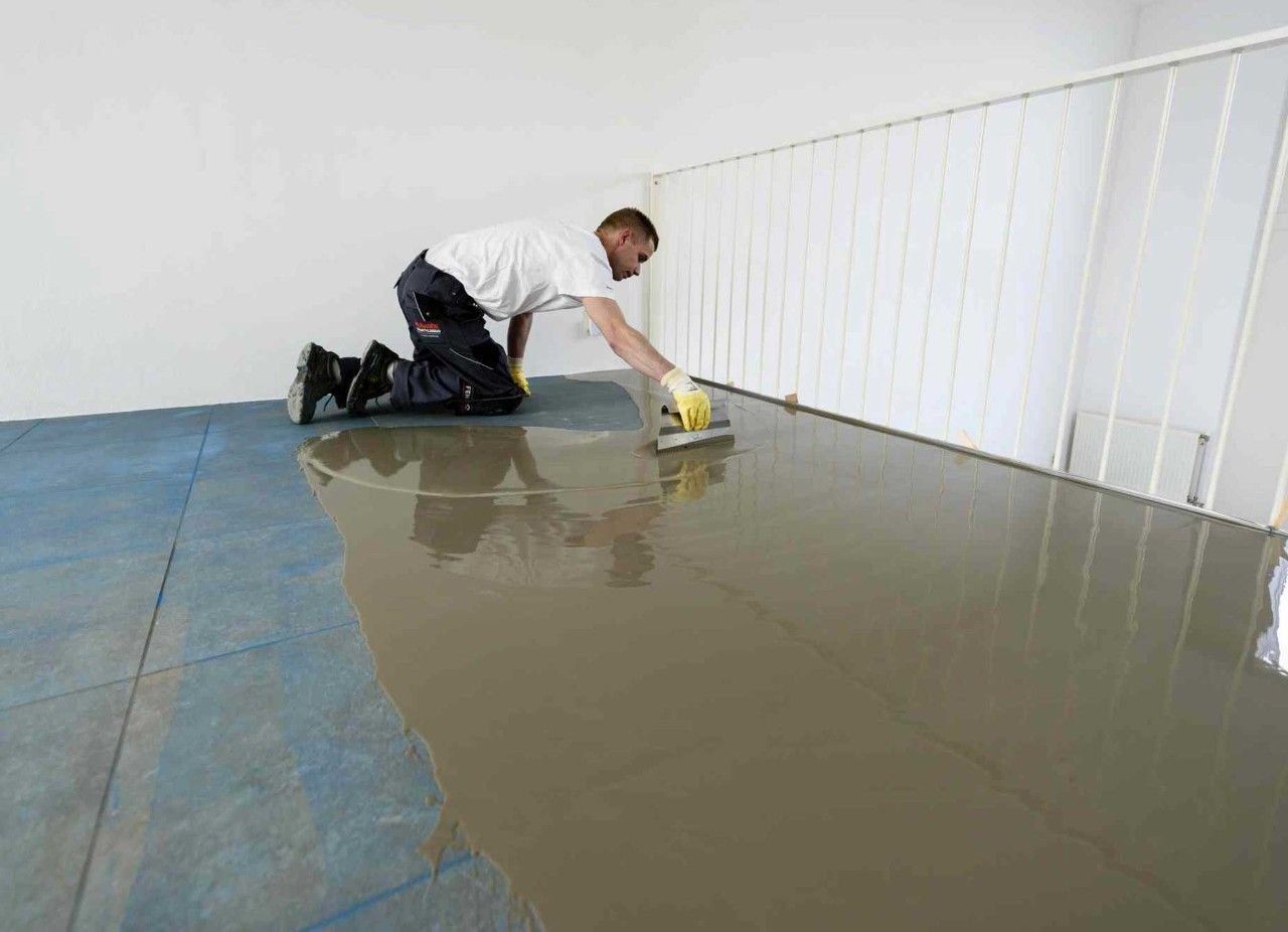 Man kneeling, smoothing wet, gray flooring with a trowel, in a white-walled room.
