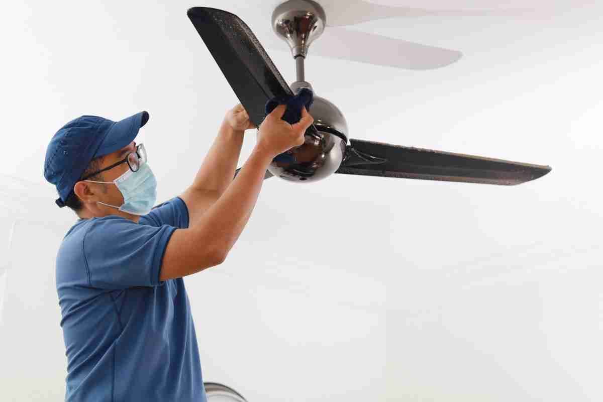 Person in blue cleaning a dark ceiling fan with mask on. White room.