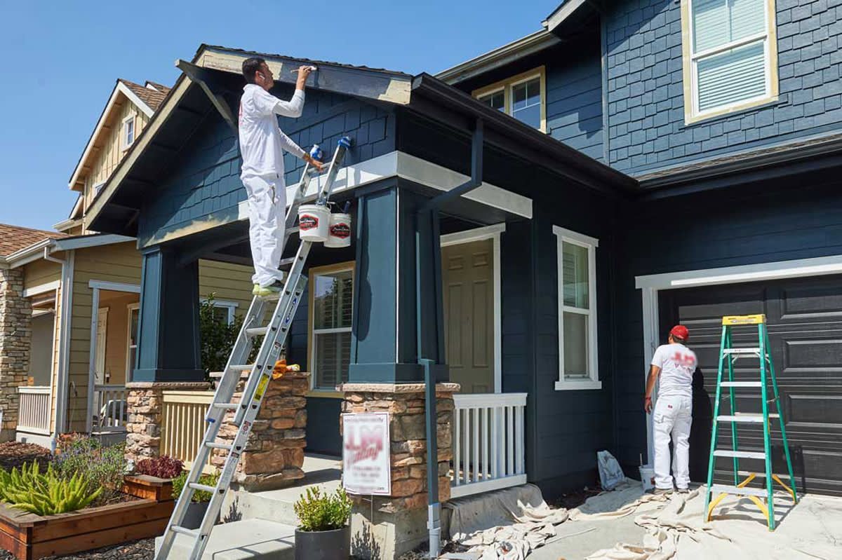 Two painters painting a dark blue house exterior. One is on a ladder, the other on a green ladder on the ground.