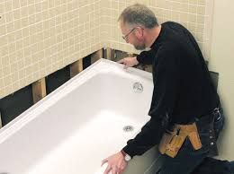 Man installing a white bathtub in a bathroom, near tile and exposed wall studs.