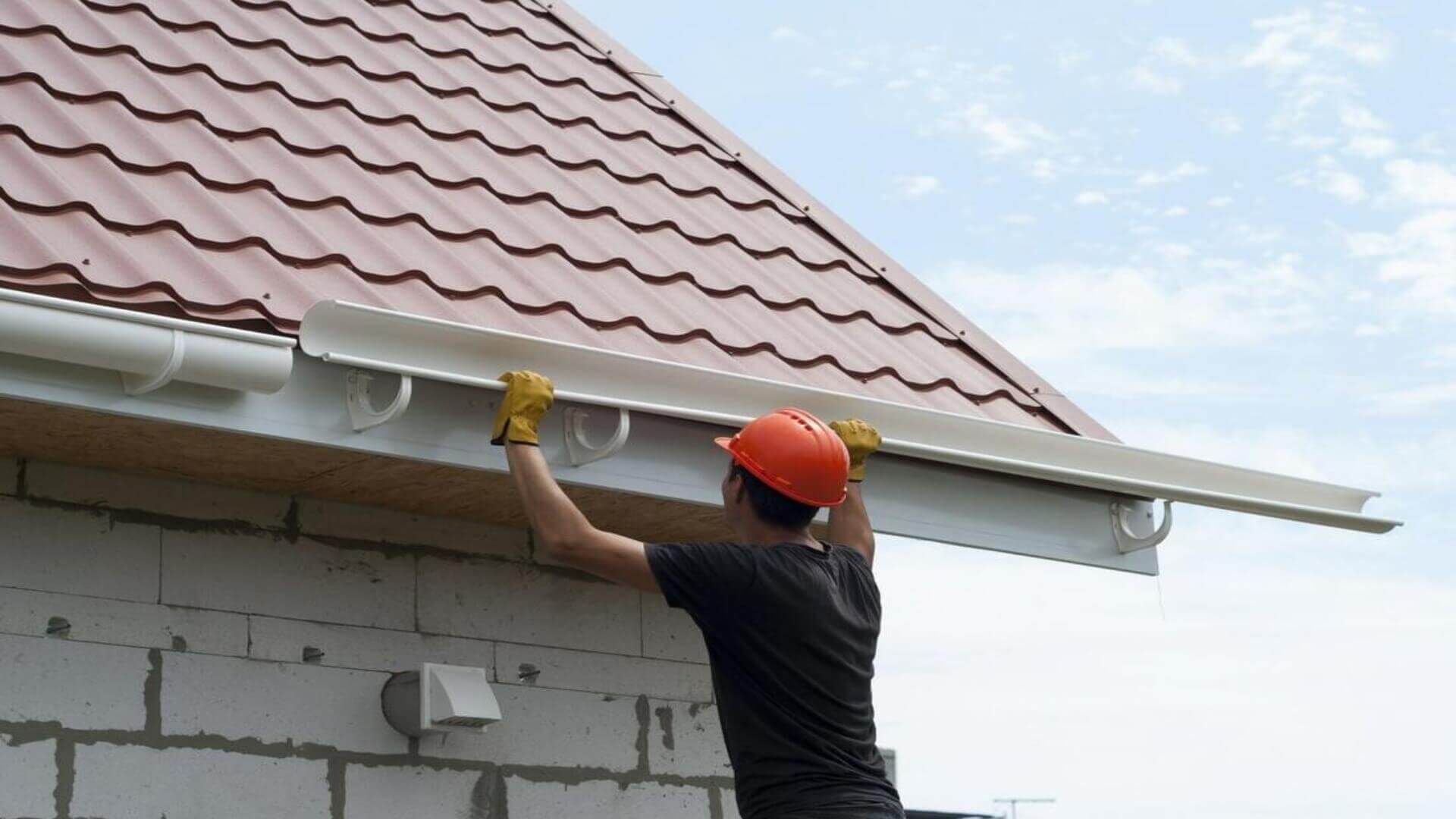 Person in hard hat installing a white gutter on a red metal roof.