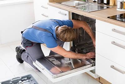 Person repairing a dishwasher in a kitchen, wearing a blue shirt and grey pants.
