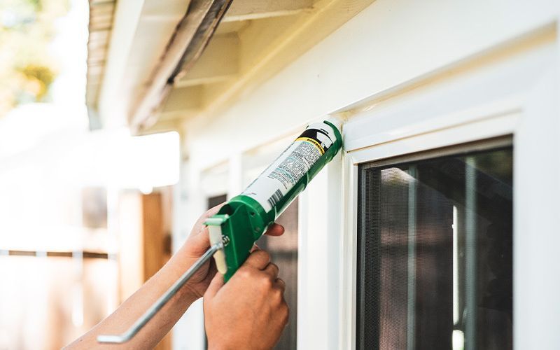 Person caulking around a white window frame with a green caulking gun.
