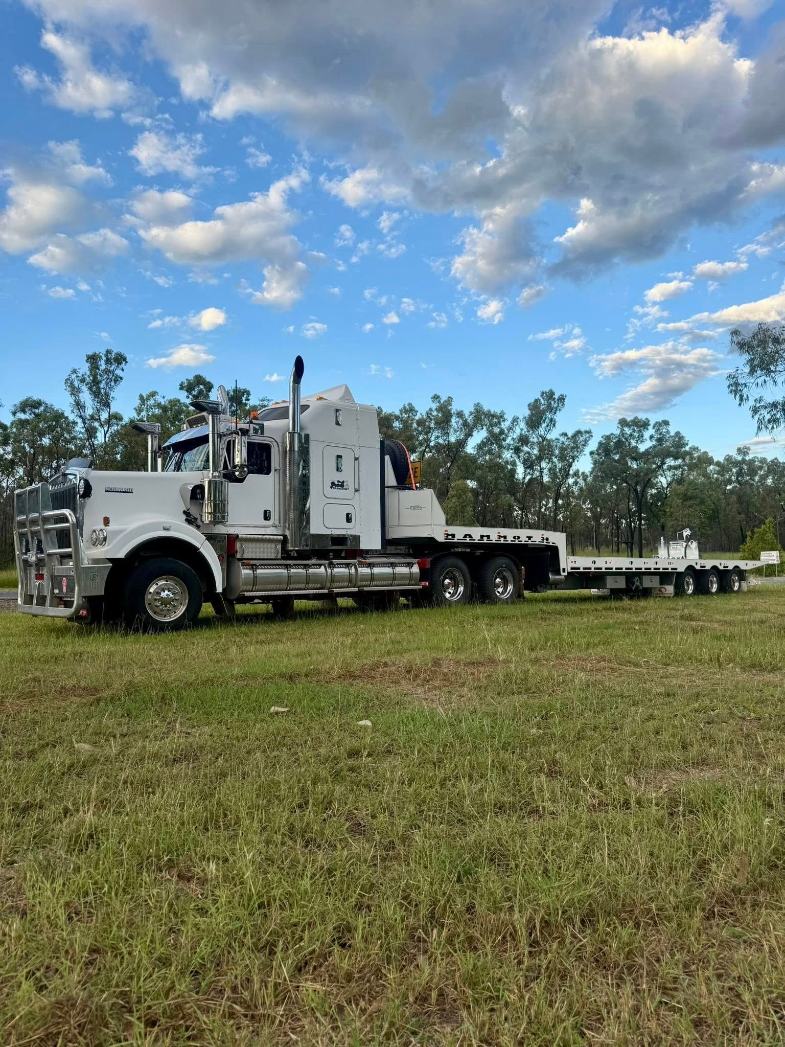 White utility truck with flatbed trailer parked in a grassy field under a cloudy blue sky — Full Torque Transport In Parkhurst, QLD 