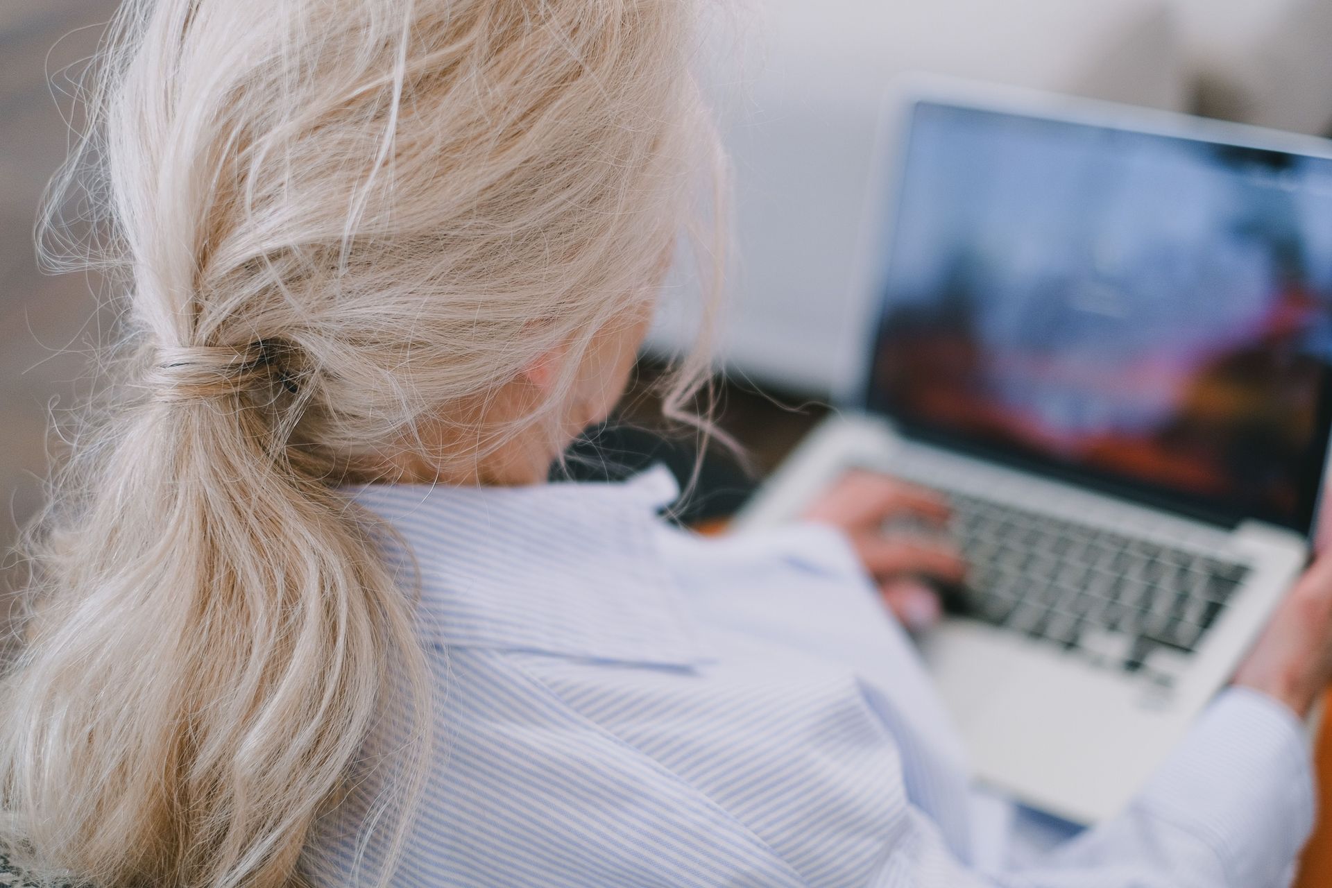 Person using a laptop outdoors, seen from behind, with long blonde hair and a light striped shirt