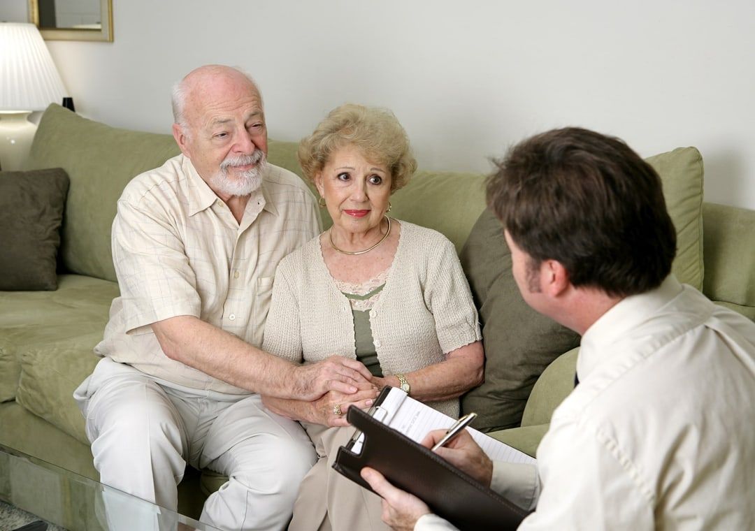 An elderly couple is sitting on a couch talking to a man holding a clipboard.