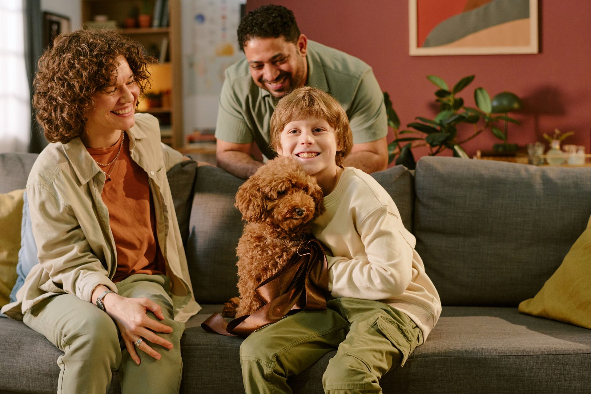 Family smiles on couch with a brown dog. A man stands behind them, all in a cozy living room.