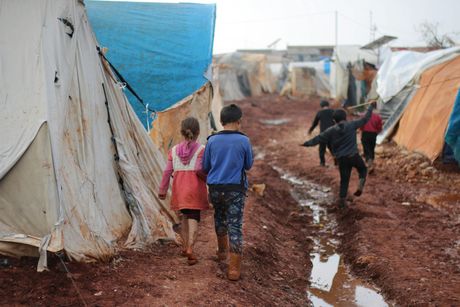 A group of children are walking through a muddy area in front of tents.