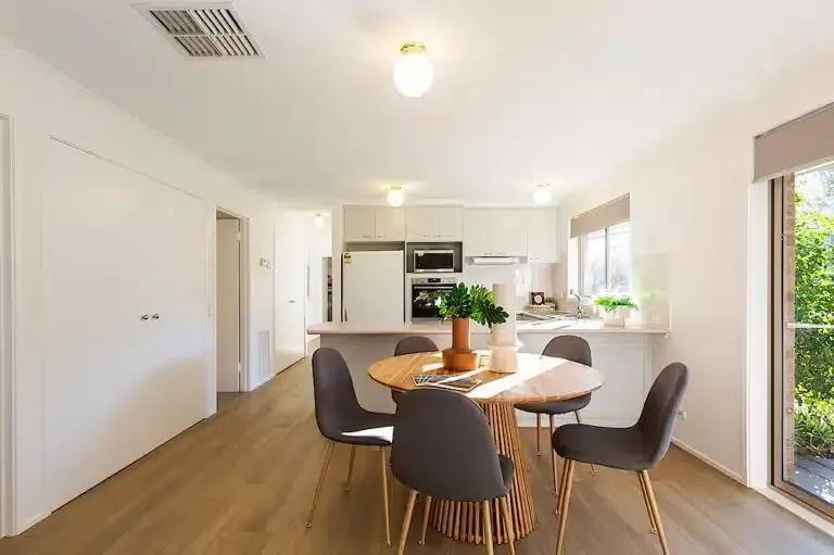 Dining area with round wooden table, gray chairs, and kitchen in background; bright, sunny interior.
