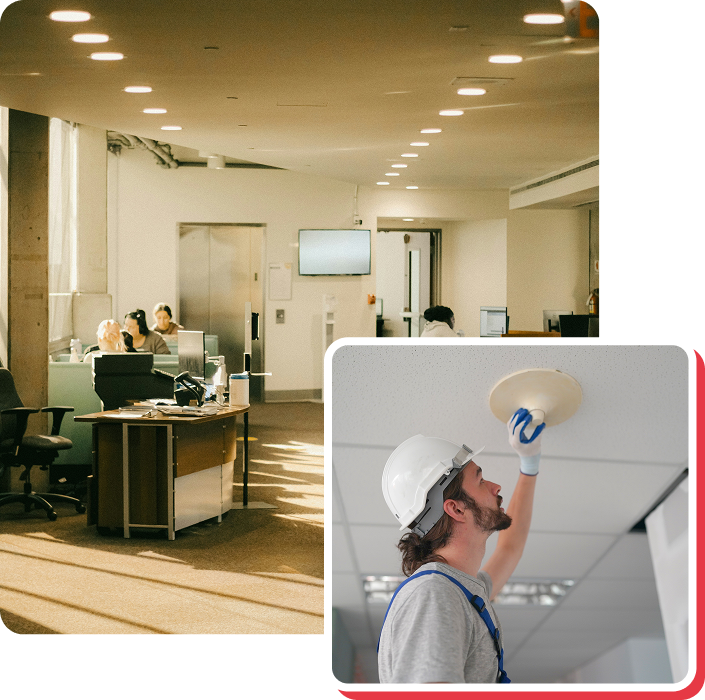 An electrician installing a ceiling light in an office. A person is sitting at their desk in the background.