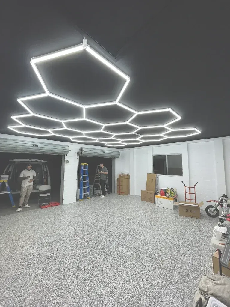 Garage with hexagonal LED lights, people, boxes, and a scooter. Black ceiling, speckled floor, and open garage door.