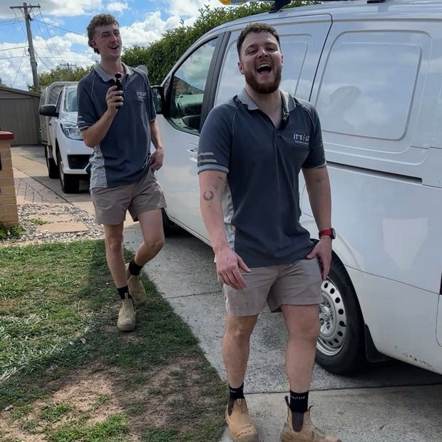 Two people wearing grey work shirts and khaki shorts laughing in front of a white van parked on a residential driveway.
