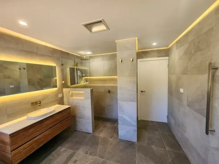 Modern bathroom with gray tile, wooden vanity, and illuminated mirrors and accents.