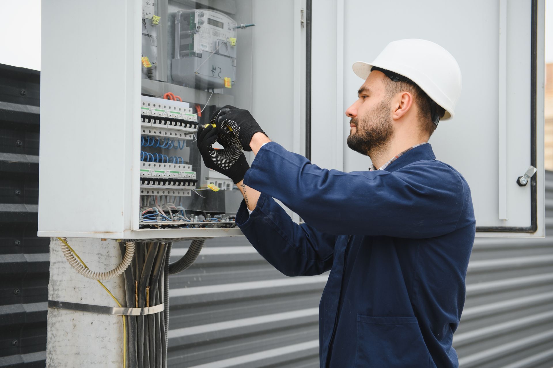Electrician in white helmet, blue uniform, working on a gray electrical panel outdoors.