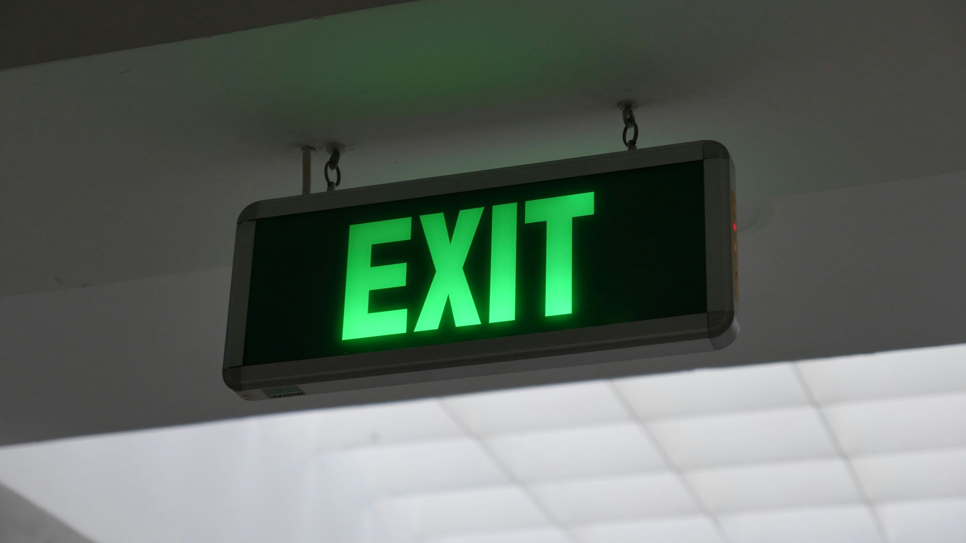 A glowing green EXIT sign hanging from a ceiling against a white, indoor background.