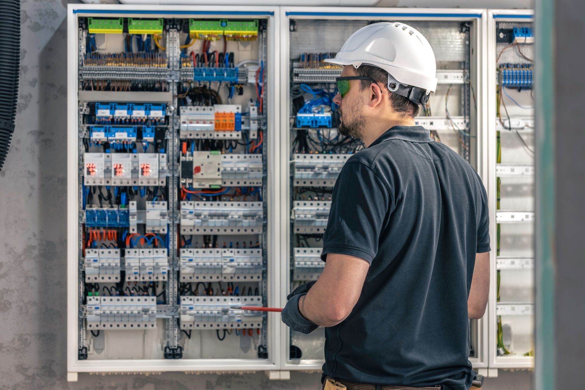 An electrician in a hard hat and safety glasses inspects a large industrial electrical panel with various components.