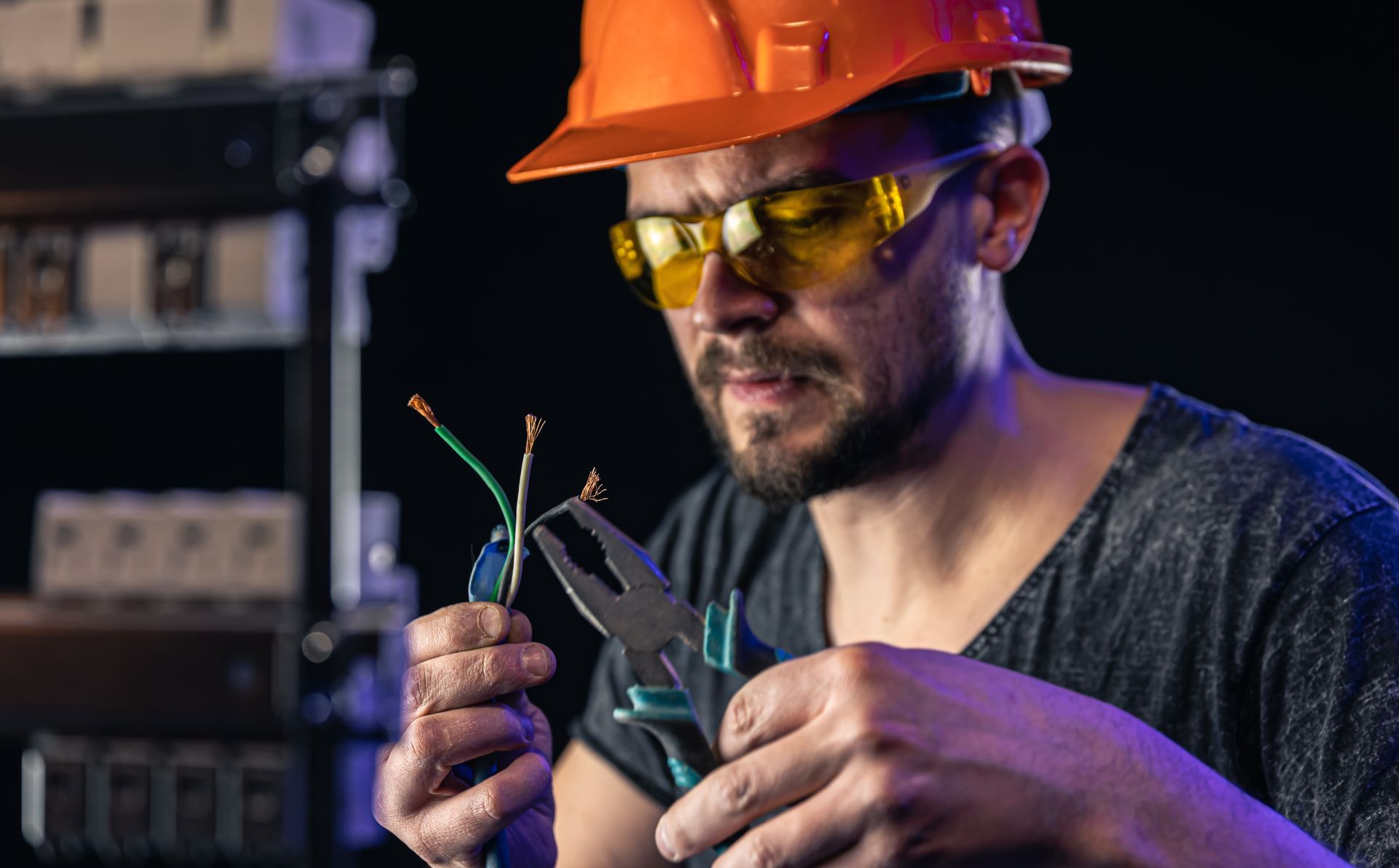 An electrician in a hard hat and safety glasses uses pliers to work on exposed electrical wires against a dark backdrop.