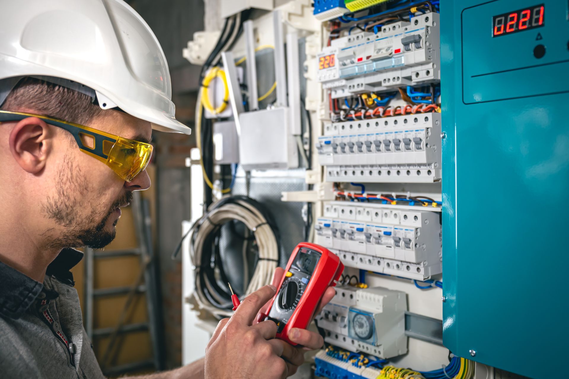 Electrician in a hard hat and yellow safety glasses using a red multimeter to test a circuit breaker panel.