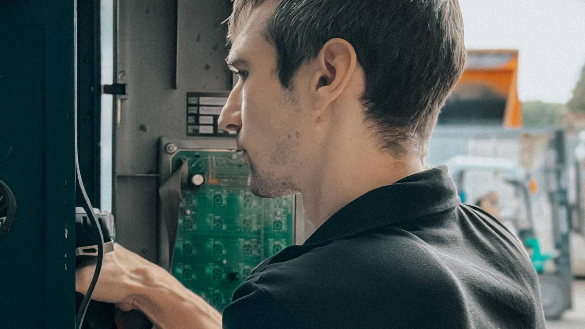 Man in black shirt at a machine, turning a dial. Outdoors, green and gray tones.