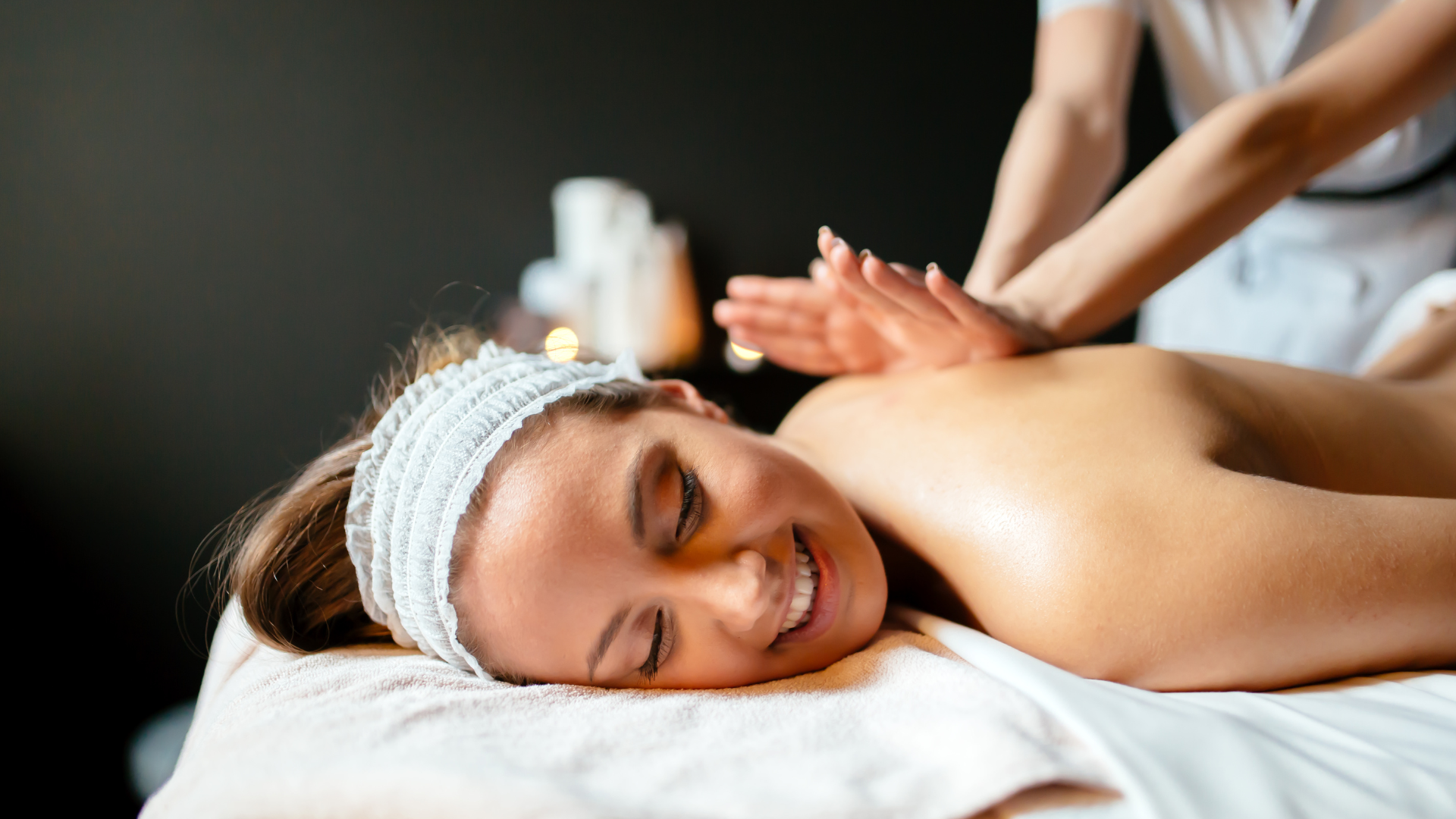 Woman receiving a back massage on a spa table, smiling with a white towel headband.