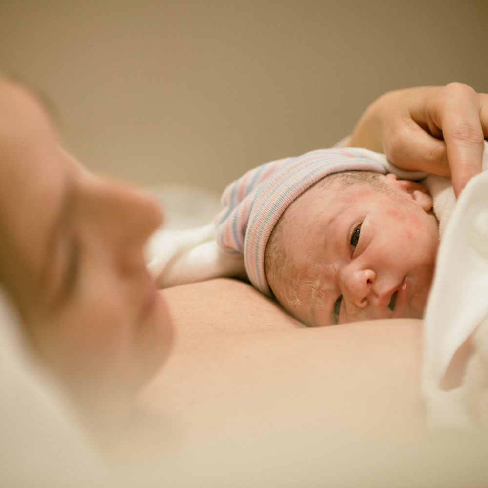 Newborn wrapped in a blanket, resting on a parent’s chest, with a hand gently holding the baby.