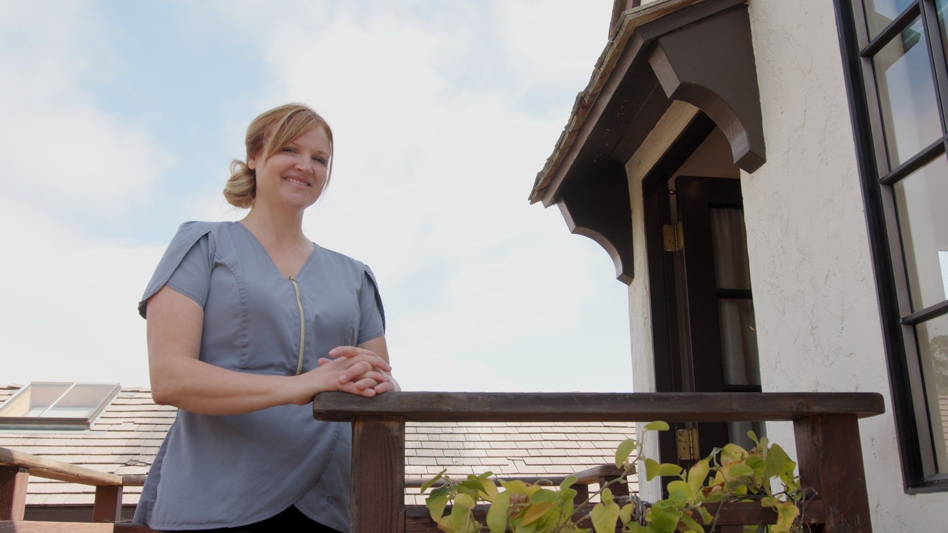 Person leaning on a wooden railing outside a building, wearing a light blue blouse under a bright sky