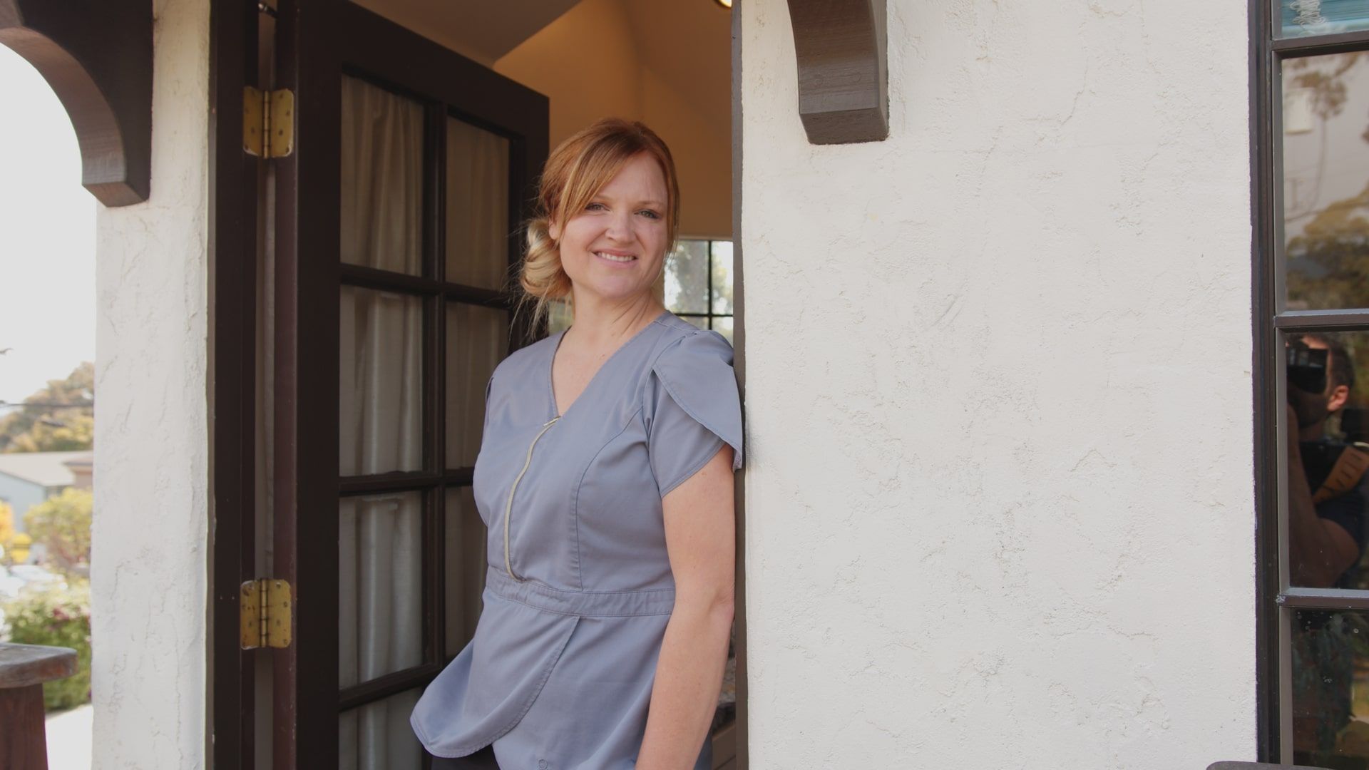 Woman standing in a doorway beside a white wall, wearing a blue blouse and smiling.