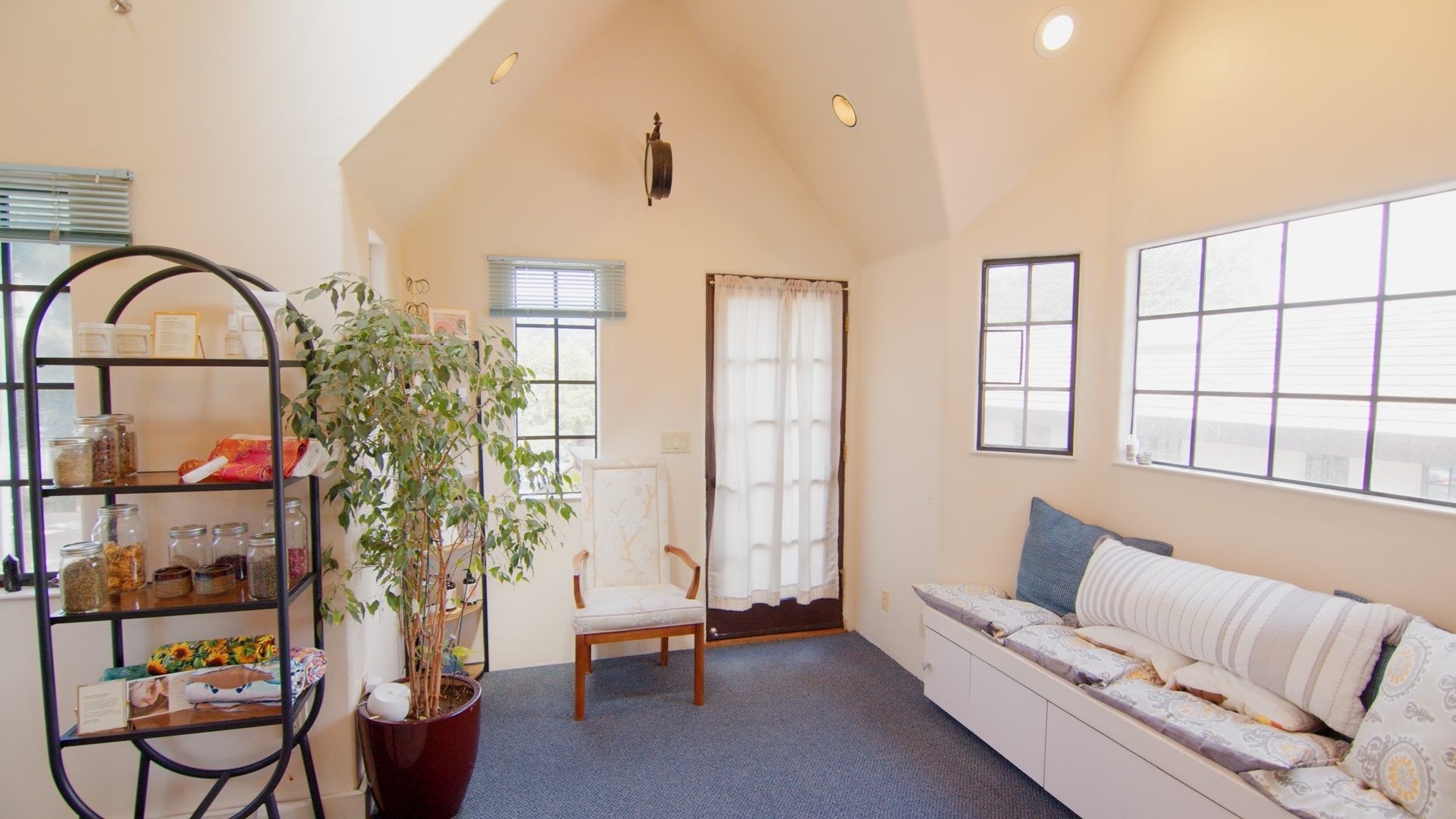 Bright living room with beige walls, large windows, a white daybed, and a potted tree on a shelf.