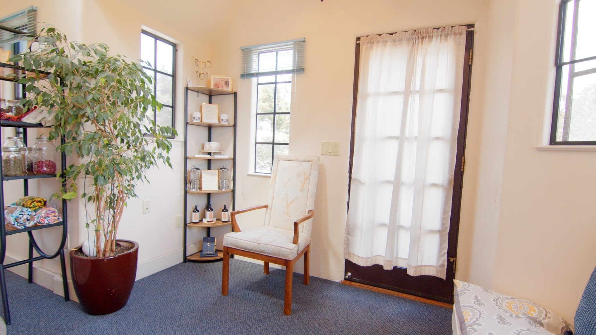 Bright sunroom with a white chair, potted plant, shelves, and sheer curtains by tall windows.