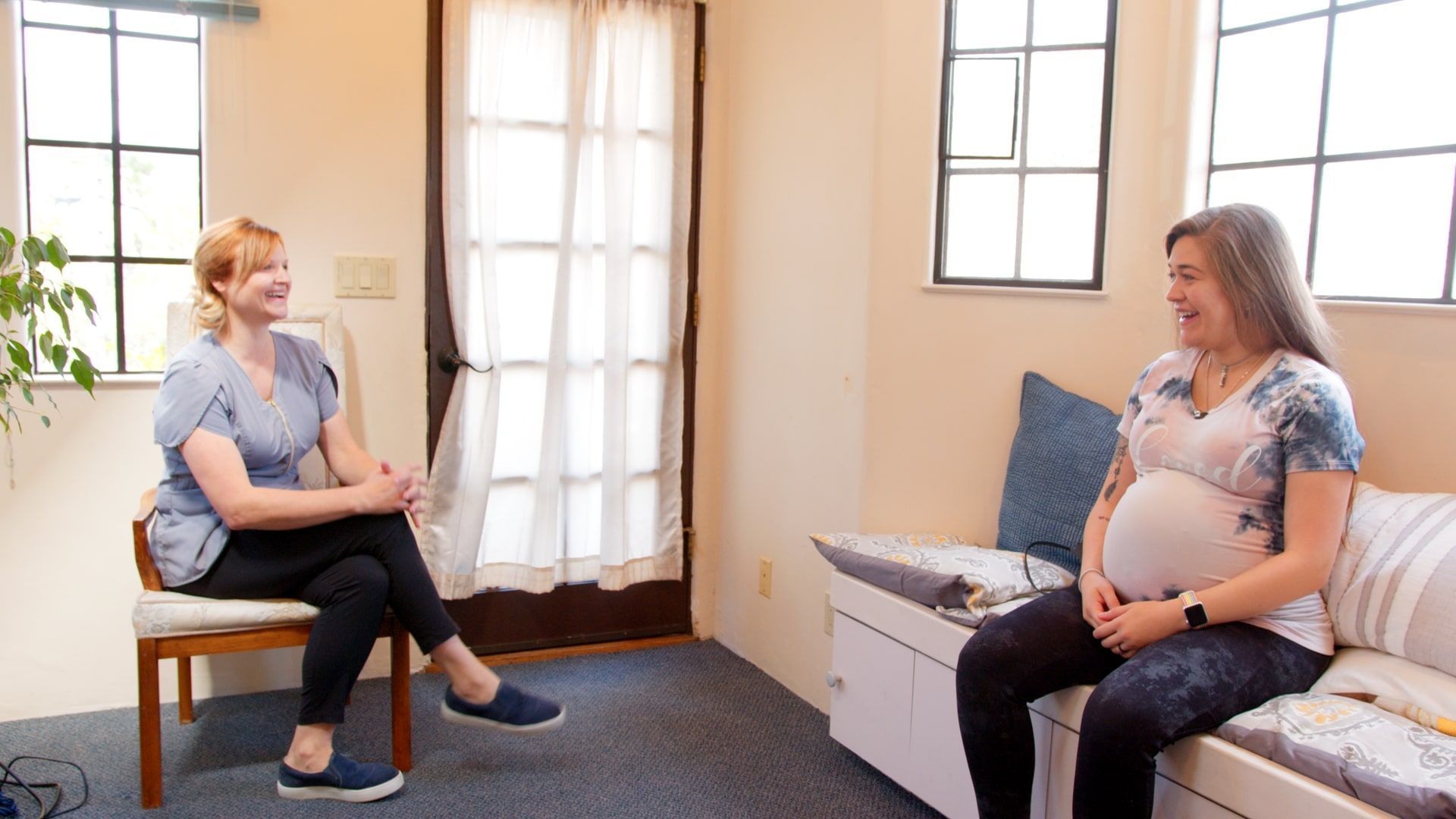 Two women sit facing each other in a bright living room, one visibly pregnant, in conversation.