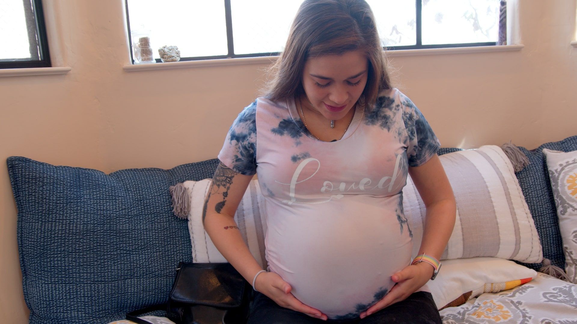 Pregnant person in a pink shirt sitting on a couch, smiling and holding their belly.